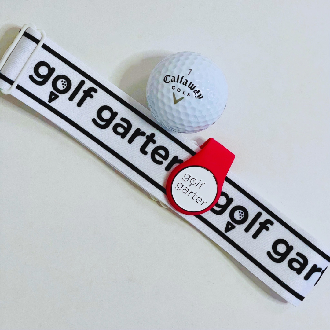 This is a photograph of a berry coloured band with a golf garter branded ball marker wrapped over a Golf Garter. There is a golf ball beside it. 