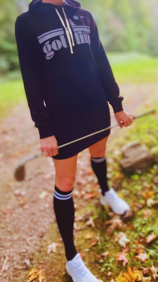 This is a photograph of a woman on the golf course. She is wearing a black hooded sweatshirt dress. It features a bold design that says Golf Life. She's wearing black knee-high socks and holding a golf club.