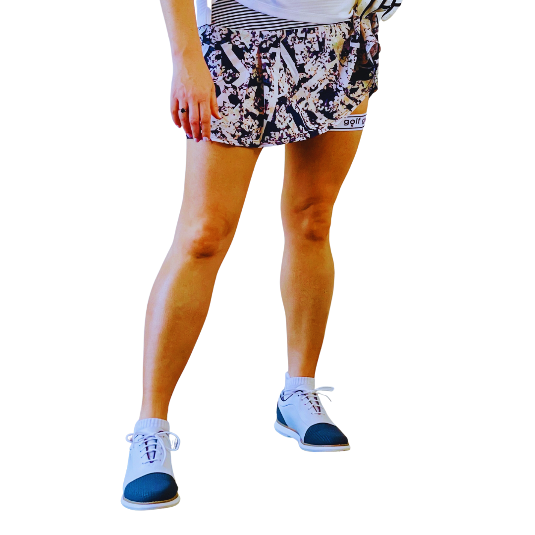 Photograph of the lower half of a woman's body. The woman is wearing a multi-colored golf skirt and golf shoes. On one hand, the woman is wearing a golf glove. She is lifting part of her skirt to show a Golf Garter on her left thigh.